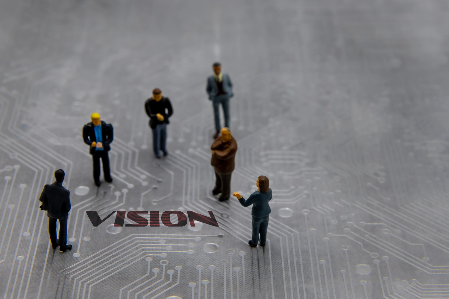 6 Miniaturfiguren aus Plastik, wie man sie von Modelleisenbahnen kennt, stehen in loser Aufstellung auf einer Halbleiterplatine. In die Halbleiter-Maserung ist ein Wort abgedruckt: Vision. Die Figuren, 5 Männer und eine Frau stehen um dieses Wort herum, blicken zu ihren Füßen auf das Wort und scheinen nachzudenken und im Dialog ihre Vision abzuleiten.