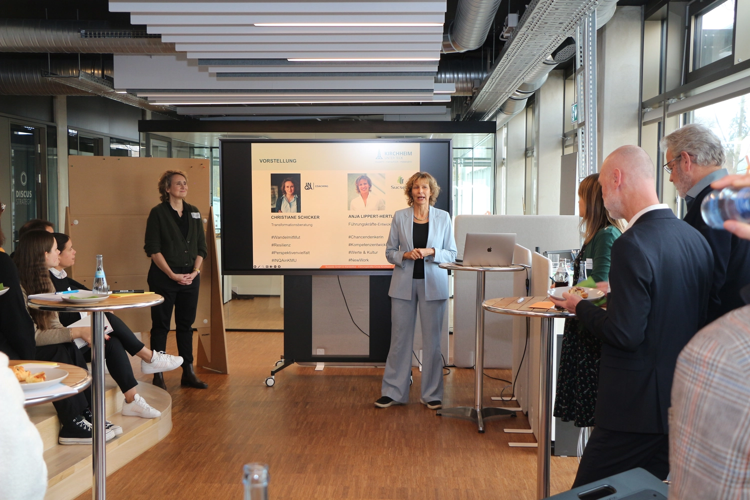 Führungskräfte Circle 2024-26 - Christiane Schicker steht links neben einem großen Monitor, Anja Lippert-Hertle steht am rechten Rand des Monitors. ALH moderiert in diesem Moment, dem Publikum frontal zugewandt, während CS zuhört. Die Veranstaltung findet in einem Loft-Büro statt, im Vordergrund und an der Seite sieht man Teilnehmer an Stehtischen und auf Treppenstufen sitzend, die der Moderation folgen. Die Teilnehmer haben teilweise kleine Teller mit Snacks in der Hand, auf den Stehtischen stehen Gläser und Getränkeflaschen - die Veranstaltung fand mittags bei Tageslicht statt, dass durch die seitlicher Fensterfront am rechten Bildrand den Raum erhellt. Auf dem Monitor sieht man Portraitbilder von CS und ALH mit der Überschrift "Vorstellung". CS und ALH präsentieren sich auf dem Bildschirm mit jeweils 4 Schlüsselworten, die ihre Tätigkeit zusammenfassen. Bei CS stehen die hashtags: #WandelmitMut #Resilienz #Perspektivenvielfalt #INQAmitKMU. Bei ALH sind folgende hashtags zu lesen: #Chancendenkerin #Kompetenzentwicklung #Werte&Kultur #NewWork.