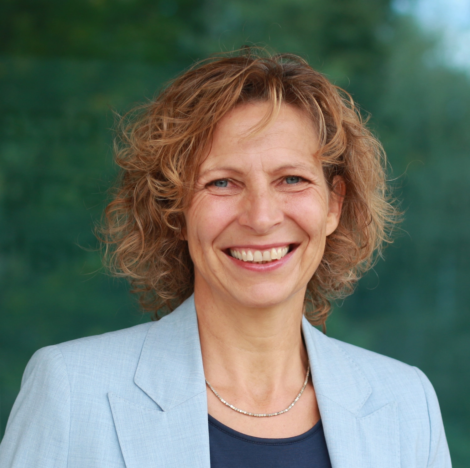 Portraitbild Anja Lippert-Hertle - Anja blickt mit fröhlichem Lachen entspannt und strahlend frontal in die Kamera. Sie trägt ein hellblaues Jackett mit einem blauen Rundhals-Tshirt. Im Hintergrund sieht man sommerlich belaubte Hecken und einen Zipfel blauen Himmel.