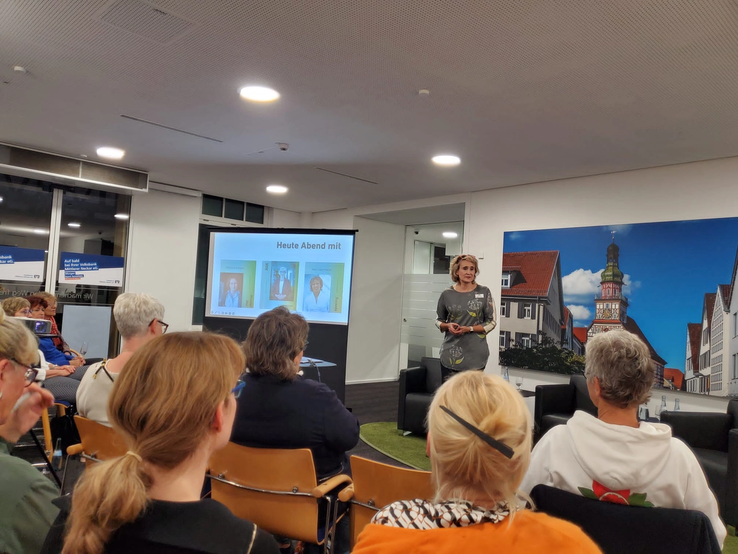 Eine Veranstaltung mit gutbesuchtem, meist weiblichen Publikum zu den Frauenwirtschaftstagen Baden-Württemberg in der Volksbank Kirchheim unter Teck. Christiane Schicker steht auf dem Podium und spricht vor dem Publikum. Im Hintergrund sieht man ein Lichtposter mit dem Rathaus und Marktplatz von Kirchheim unter Teck. Links neben CS steht ein großer Bildschirm, auf dem drei Personen, die durch diesen Abend führen, abgebildet sind: Christiane Schicker, Bettina Lichtner, AOK und Anja Lippert-Hertle