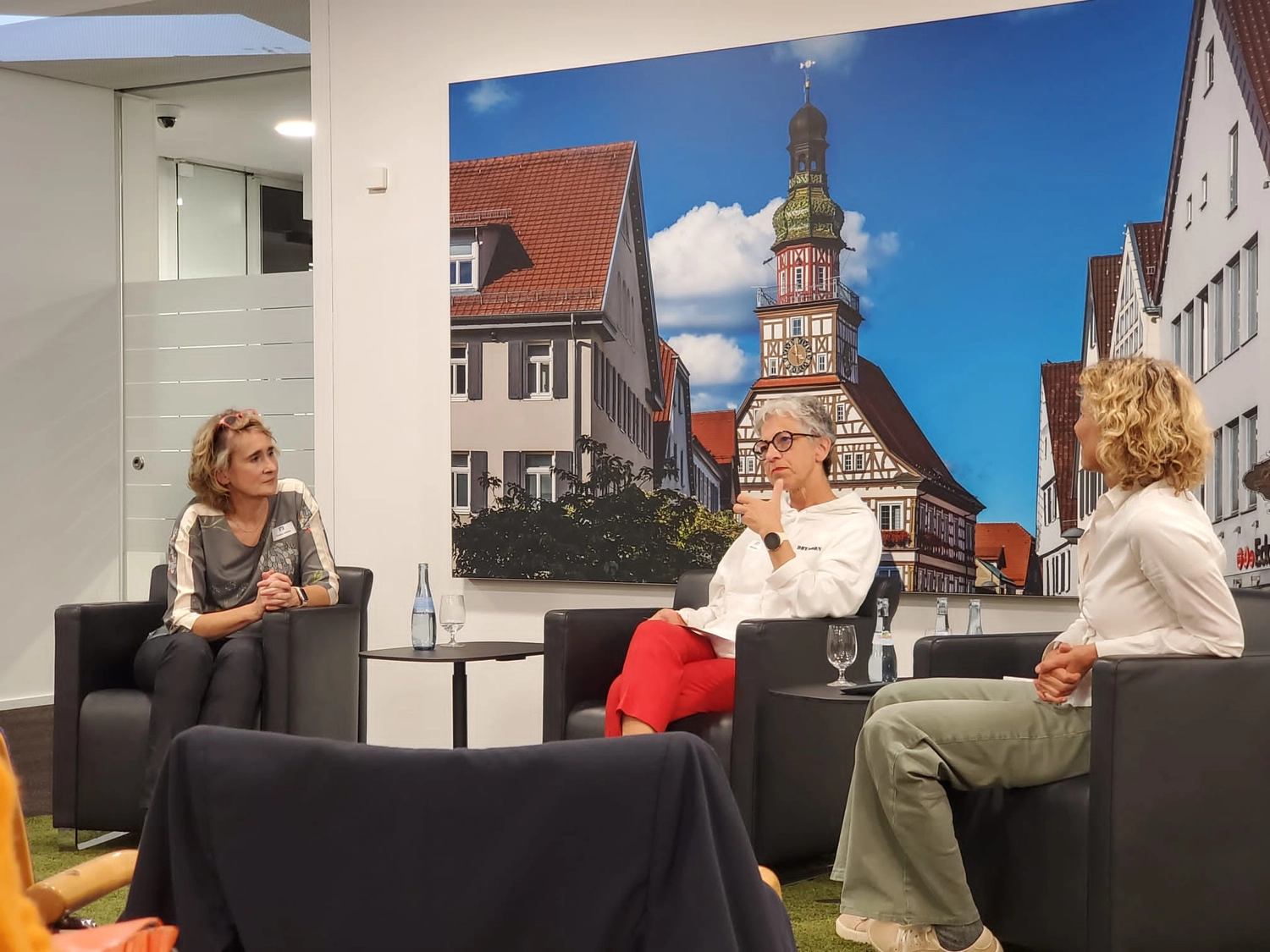 Podiumsdiskussion mit Christiane Schicker, Bettina Lichtner, AOK Baden-Württemberg, Anja Lippert-Hertle: Alle drei Personen sitzen in schwarzen Lounge-Sesseln auf einem Podium. Bettina Lichtner in der Mitte erläutert gerade etwas für das Publikum, mit Armen gestikulierend. Sie ist eingerahmt von CS und ALH, die sich ihr zuhörend zuwenden. Zwischen den Sesseln stehen kleine Beistelltische mit Getränken. Im Hintergrund sieht man das Rathaus von Kirchheim unter Teck und den Marktplatz als Lichtposter in der Schalterhalle der Volksbank Kirchheim unter Teck.