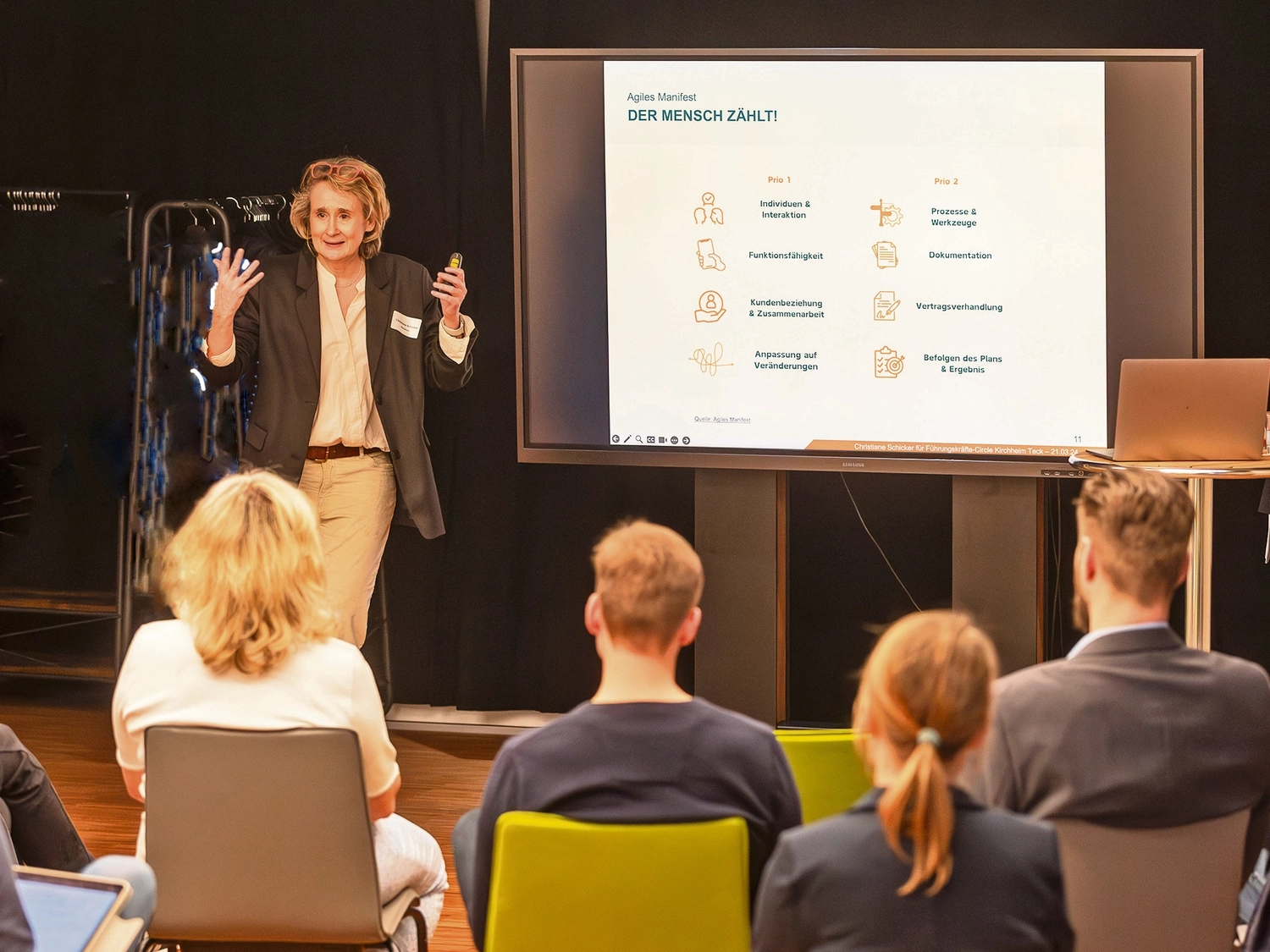 Christiane Schicker spricht von einem Podium vor einem gemischten Publikum in einem Saal. Sie hat beide Arme zur Bekräftigung ihres gesprochenen Wortes als Moderatorin gehoben. In der rechten Hand hält sie eine Fernbedienung für die Steuerung der Bildschirmpräsentation, rechts neben ihr. Das Publikum ist nur von hinten zu erkennen und sieht auf einem großen Präsentationsbildschirm rechts von CS ein Chart, von dem man die Überschrift erkennt: "Agiles Manifest - Der Mensch zählt." Rechts vom Bildschirm ist noch ein Stehpult mit dem Präsentationscomputer zu erkennen.